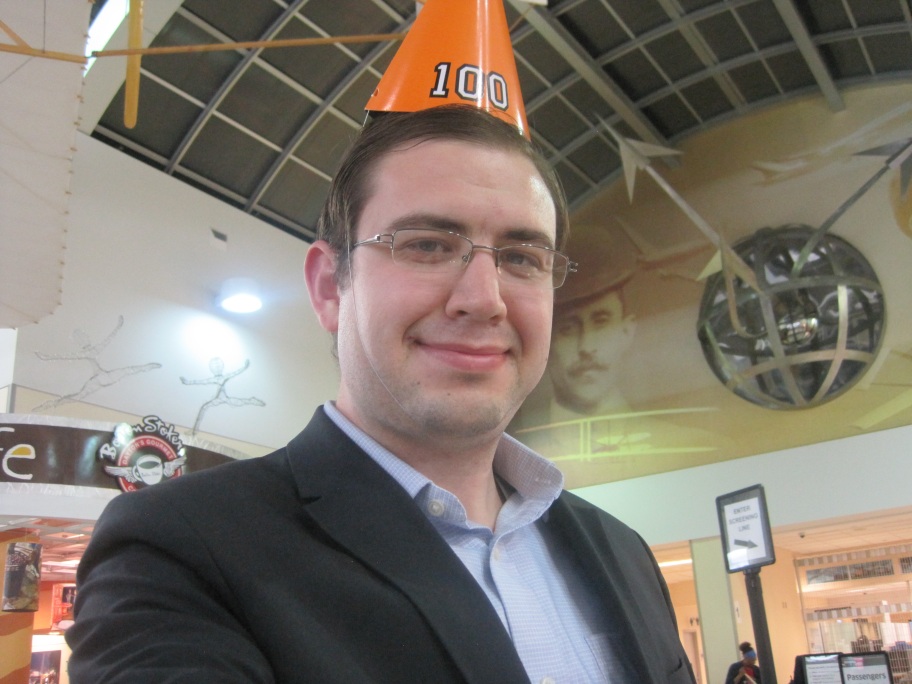 Paul, in the lobby of the Dayton airport, wearing a party hat labeled with the number 100.