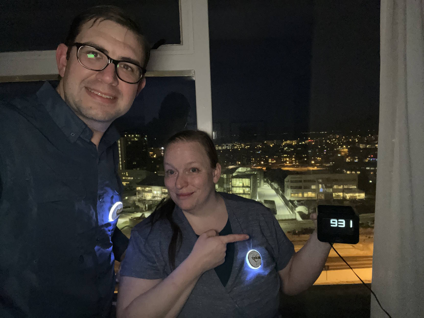 Paul and Amy holding a hotel clock that reads 9:31, in front of a window where it’s night outside.