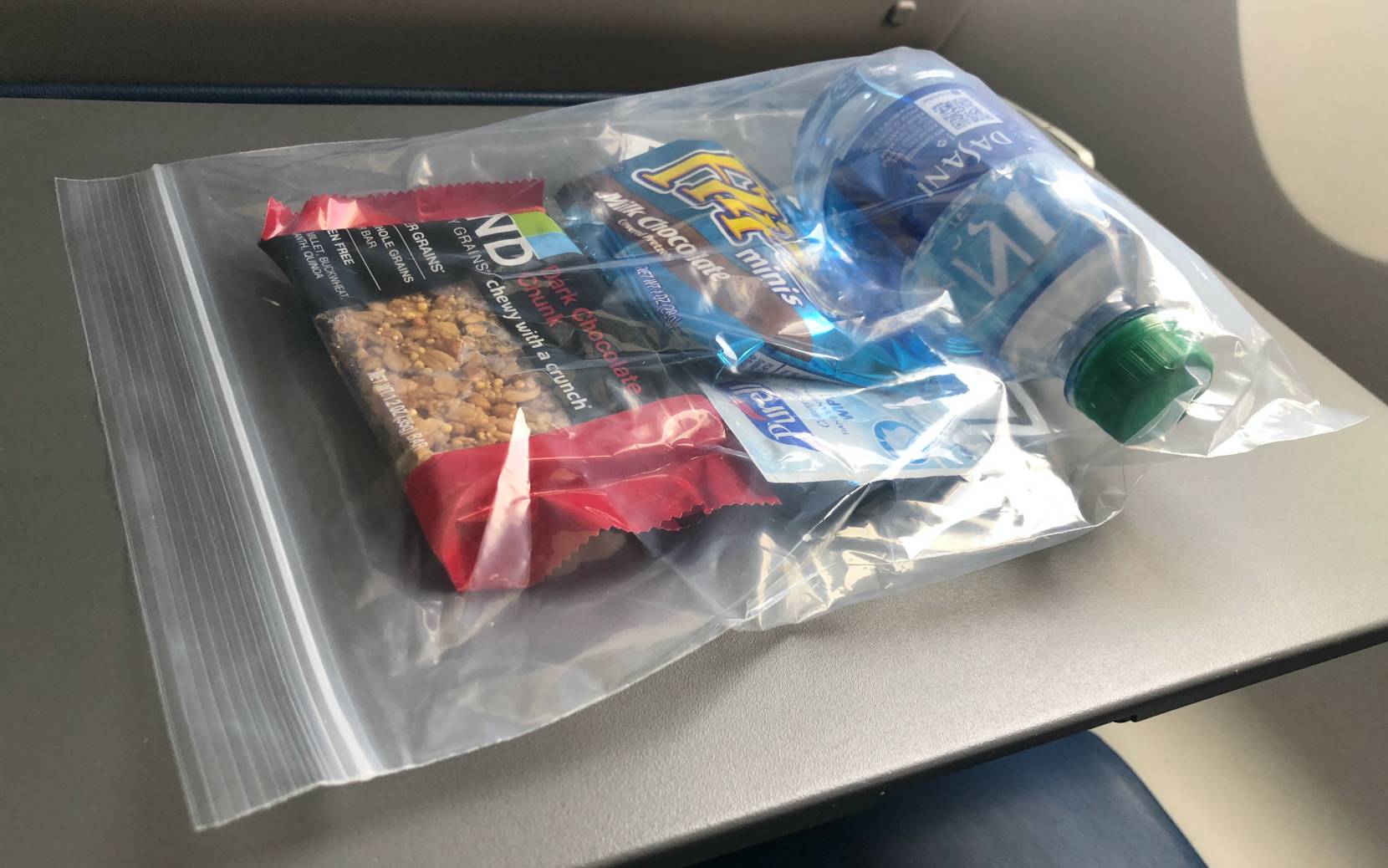 A plastic bag containing a Kind bar, chocolate-dipped pretzels, a bottle of water, and an individual hand sanitizer pack.